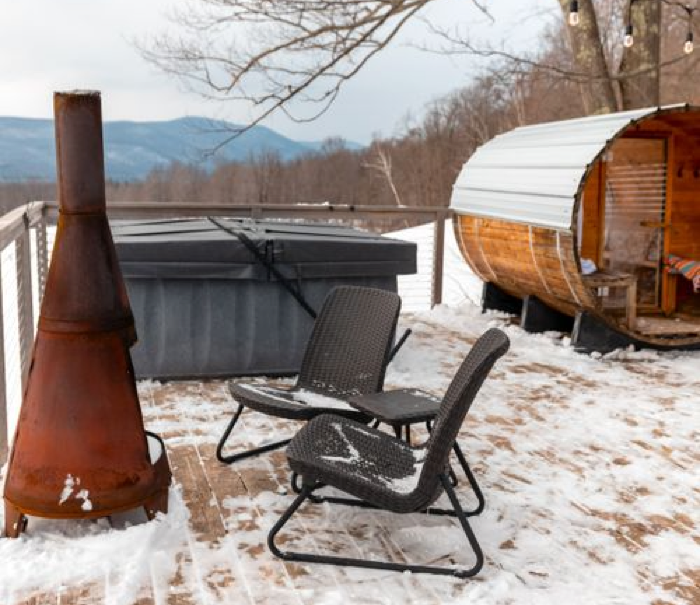 A barrel hot tub, patio heater, and chair in a snowy lakeside backyard