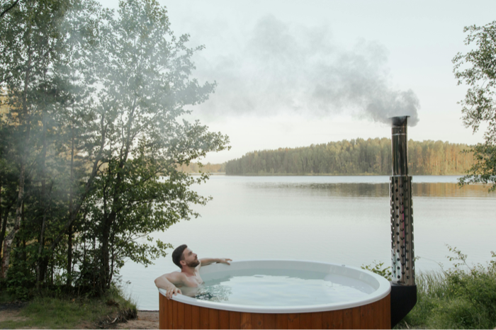A person relaxing in a wood-fired hot tub beside a calm northern lake, steam rising into the cool air
