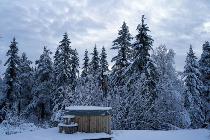 A wood-fired hot tub before a snowy evergreen forest at dusk